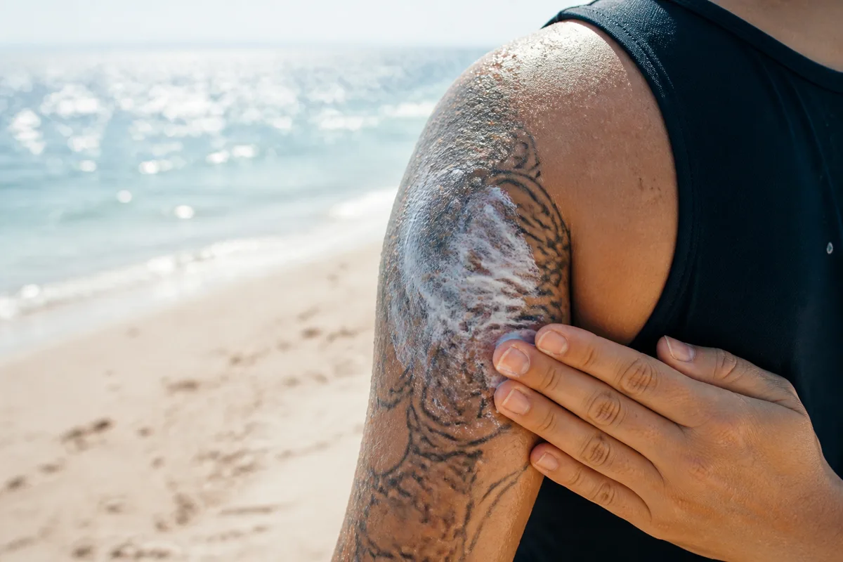 Personne appliquant une crème solaire sur un tatouage de l&rsquo;épaule au bord de la mer, lumière vive de milieu de journée