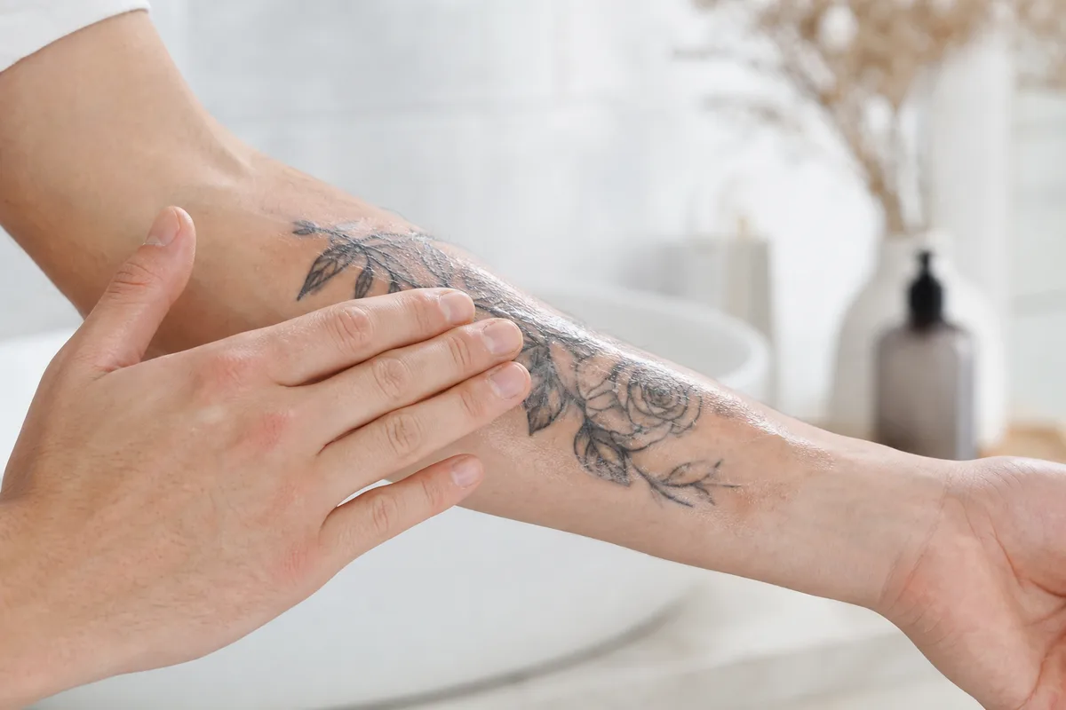 Application d’une fine couche de crème cicatrisante sur un tatouage du bras avec des mains propres, ambiance salle de bain claire